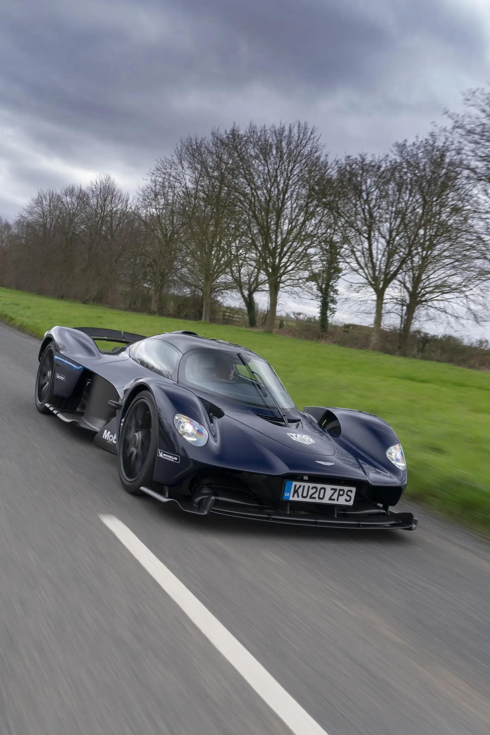 Aston Martin Valkyrie production road car in three-quarter front view on a public highway, photographed for the Aston Martin Lagonda press kit, showing the carbon-fibre body and exposed aerodynamic surfaces designed by Adrian Newey