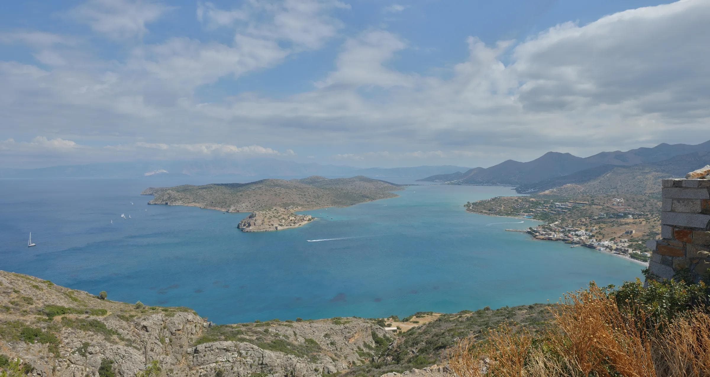 Golden-hour panorama of a Cretan hillside resort above a calm azure bay with a distant island fortress